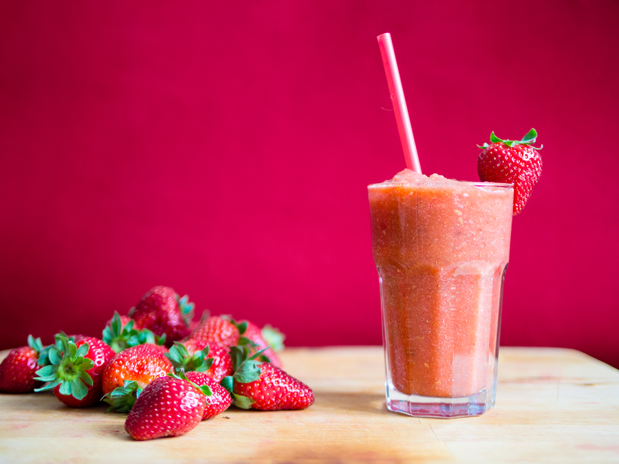 Strawberry smoothie in glass with straw and pile of strawberries with a pink background