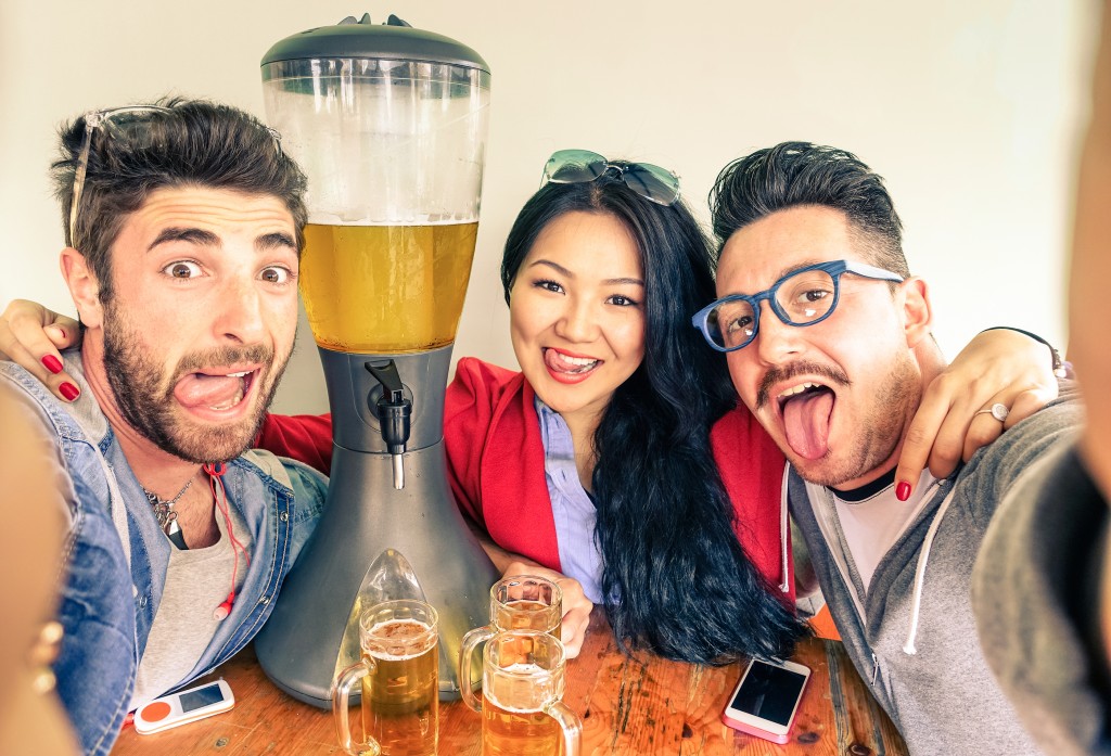 Happy friends taking selfie with funny tongue out near beer tower dispenser - Concept of friendship and fun with new trends and technology - Alternative everyday party life in vintage brewery bar