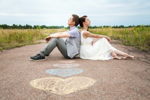wedding couple sitting on ground. love symbols on pavemant . hearts. people in love. happy bride and groom portraits. man and woman sitting back to back outside on road. newlyweds. wedding day